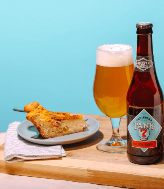 A slice of savory pie on a blue plate sits next to a glass of beer and an unopened bottle of Boulevard Tank 7 American Saison on a wooden surface with a light blue background. A folded white napkin is nearby.